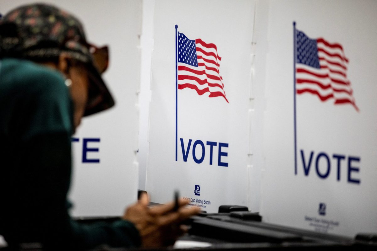 News photograph: a US polling place, voters at privacy booths beneath two American flags with VOTE printed on the panels.